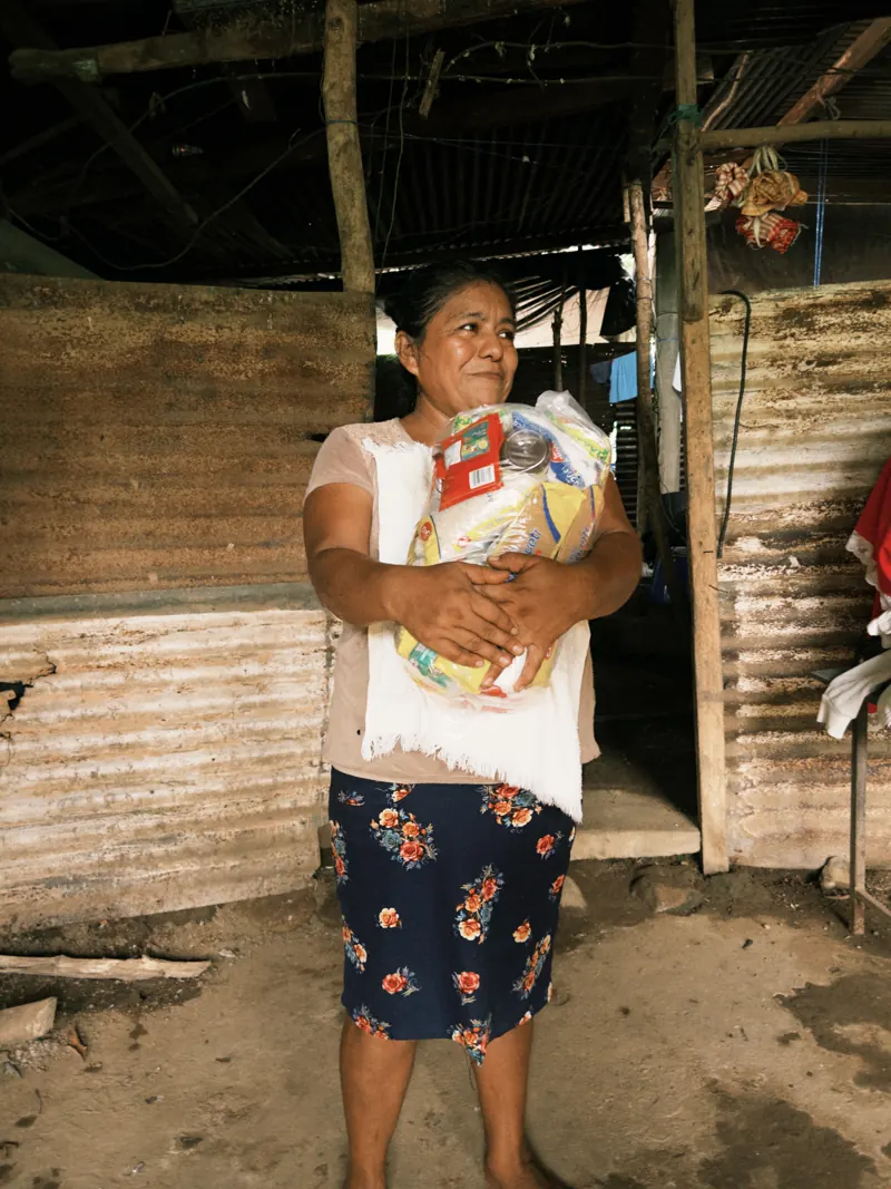 Mujer sonriendo con una bolsa de víveres frente a su casa